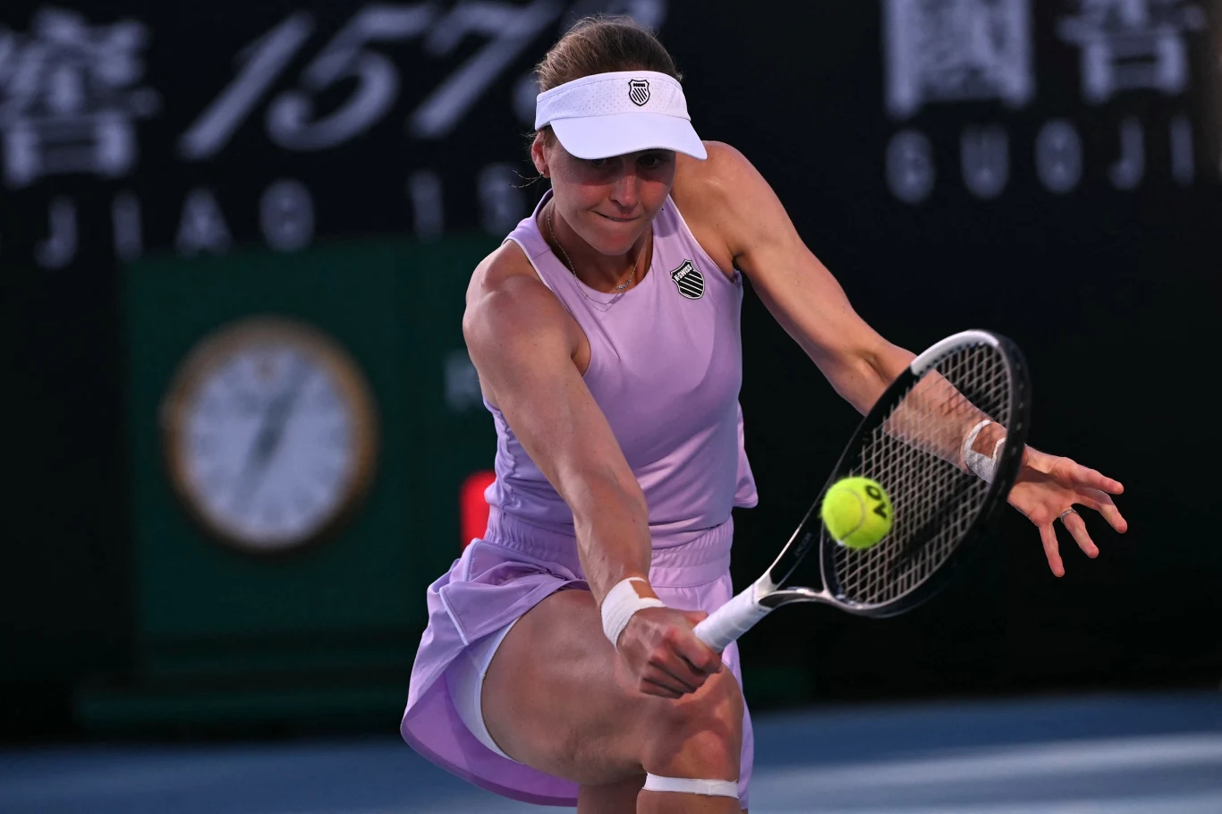 Tenisistka uderza żółtą piłkę rakietą podczas intensywnego meczu na korcie, ubrana w fioletową sukienkę sportową i białą opaskę na głowie, w tle tablica wyników i ciemne otoczenie kortu.