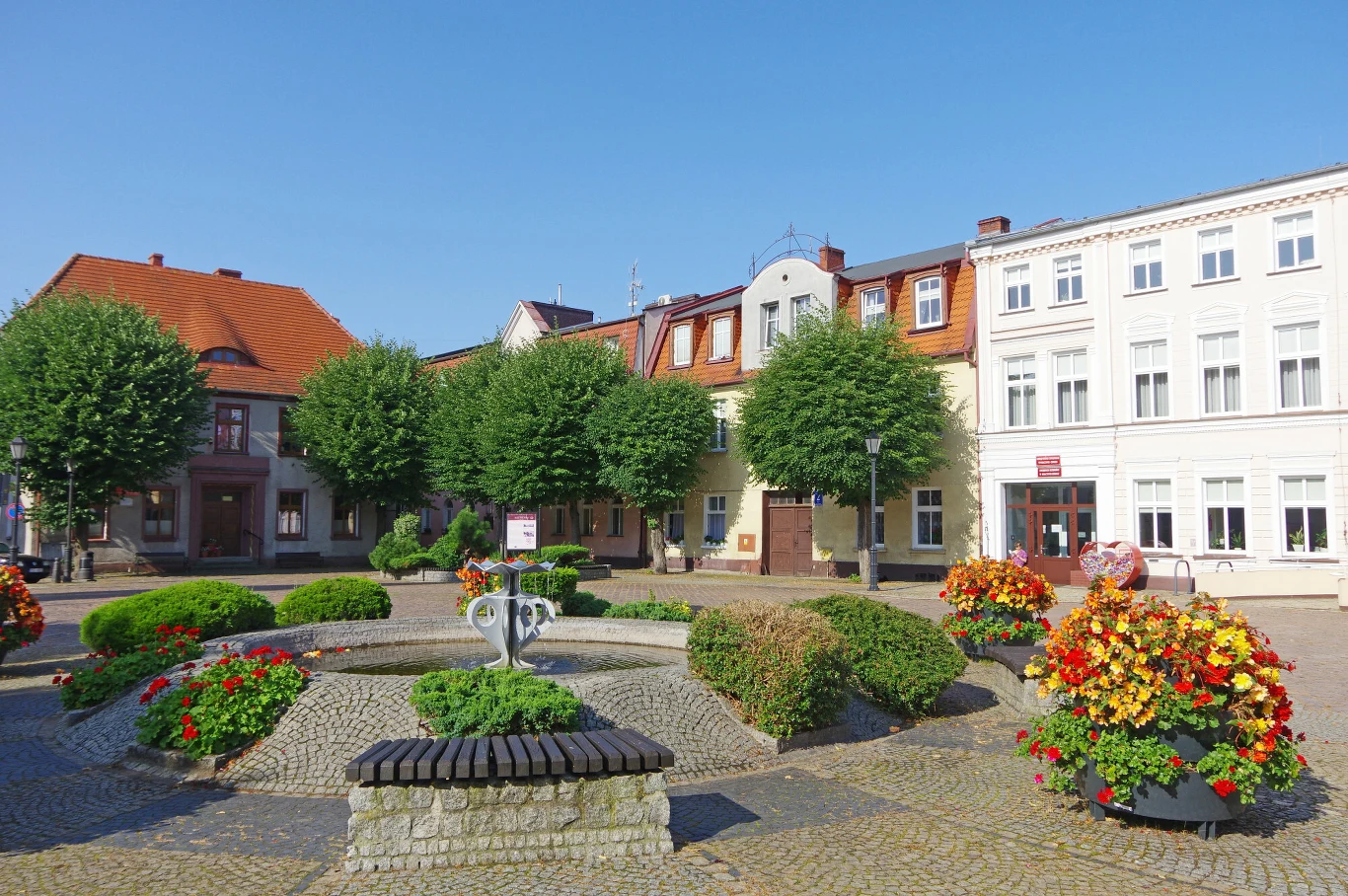 Centrum niewielkiego miasta z brukowanym placem, kwiatowymi rabatami, okrągłą fontanną pośrodku oraz zadbanymi budynkami mieszkalno-usługowymi w tle, otoczonymi zielonymi drzewami.