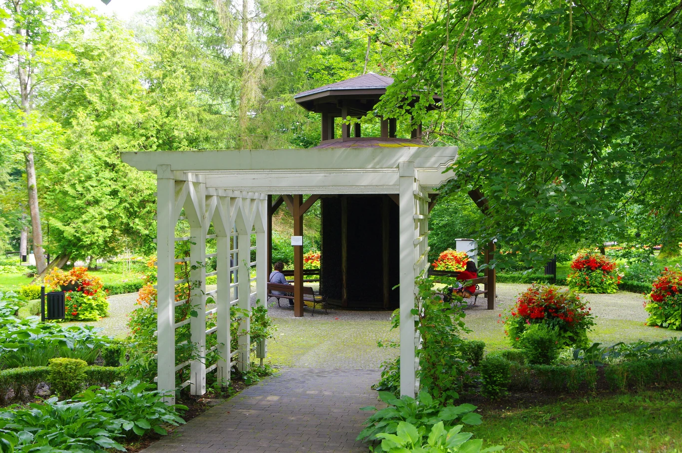 Pergola z białego drewna prowadząca do altanki w otoczeniu bujnej zieleni, krzewów i kolorowych kwiatów. W tle ławki i spokojna część parku z dekoracyjnymi kompozycjami roślinnymi.