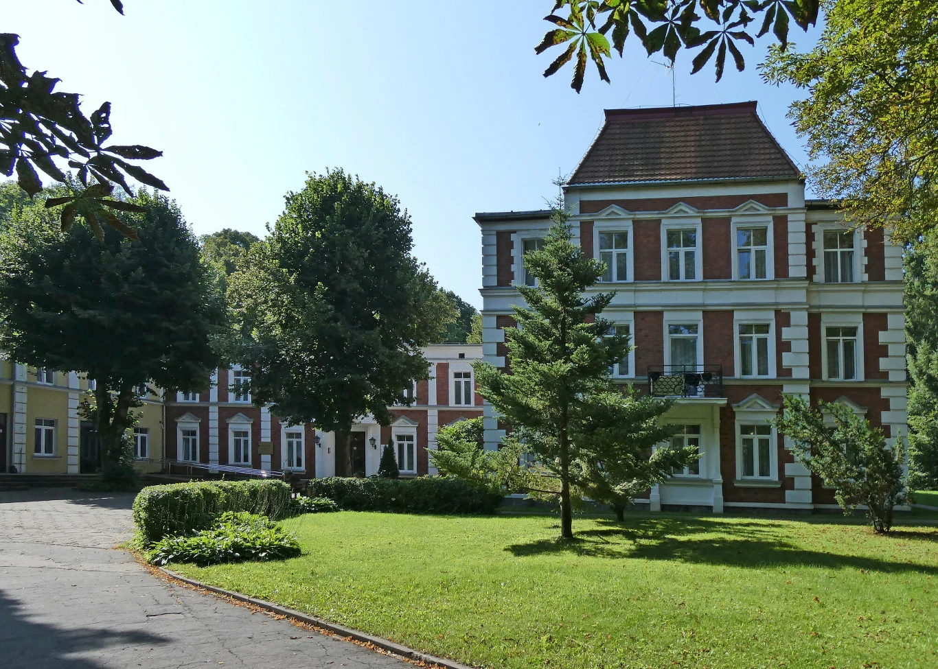 Zabytkowy budynek z czerwonej cegły z białymi zdobieniami, otoczony zadbanym trawnikiem oraz drzewami i krzewami, w tle widoczne kolejne skrzydło budynku na tle błękitnego nieba.