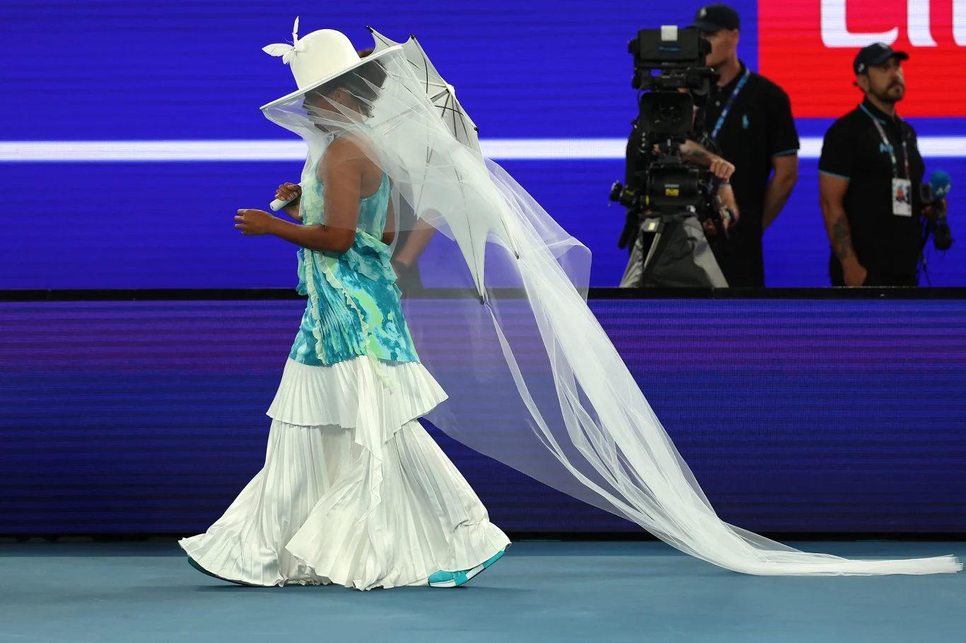 Wejście Naomi Osaki na Rod Laver Arenę Kobieta w białym kapeluszu i długim welonie ubrana w turkusową i białą suknię idzie w kierunku areny sportowej, na tle kamerzysty oraz osób pracujących przy realizacji wydarzenia, z widocznym kolorowym ekranem LED w tle.