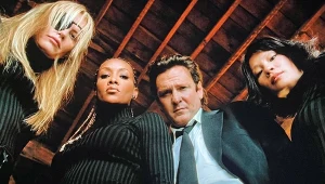 Cztery osoby w ciemnych ubraniach stoją blisko siebie, patrząc w dół w kierunku obiektywu aparatu, w tle widoczny drewniany sufit poddasza.