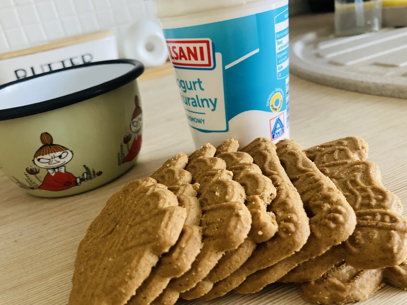 Stos herbatników ułożony na blacie obok kubka z rysunkiem postaci oraz pojemnika z jogurtem naturalnym.
