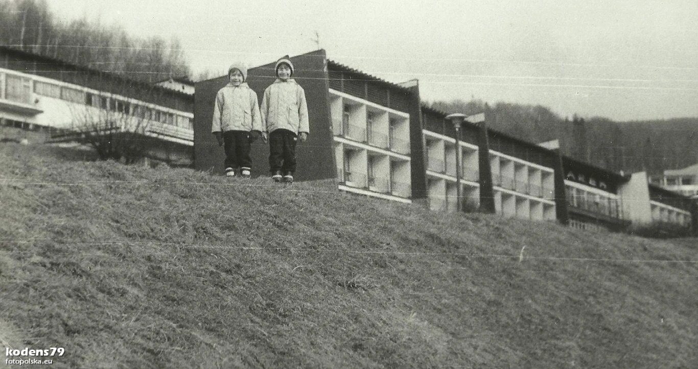 Dwójka dzieci w zimowych kurtkach stoi na stoku przed szeregiem nowoczesnych budynków na tle drzew i wzgórz. Budynki charakteryzują się dużymi oknami o regularnym układzie.