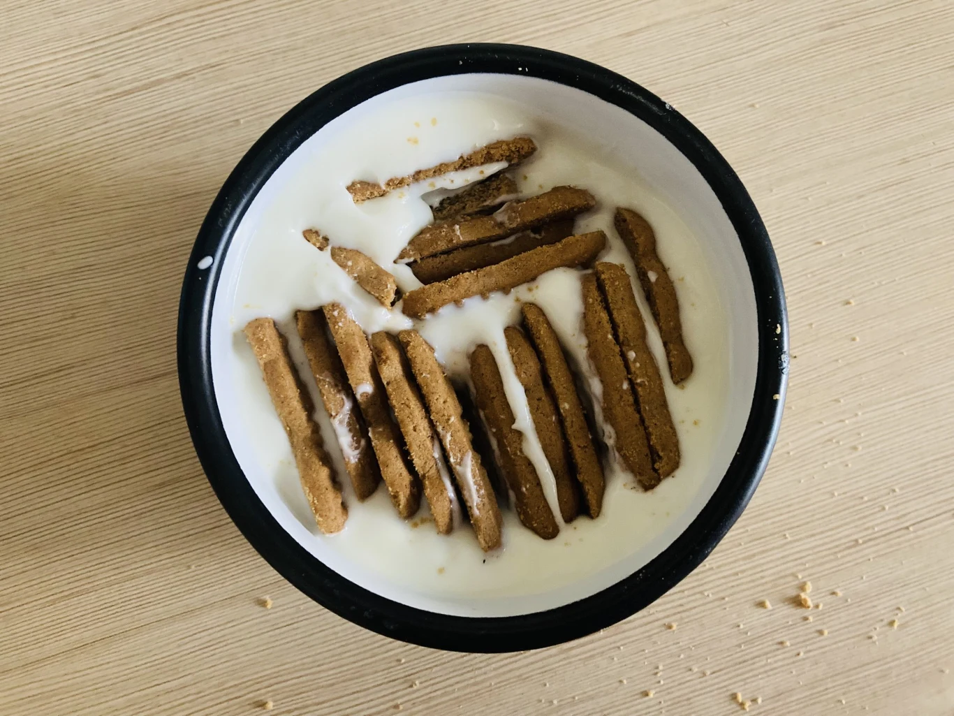 Miseczka z mlekiem, do której dodano pokruszone biszkopty lub herbatniki, leżące na mleku w nieregularnym rozmieszczeniu. Na brzegu miski widoczne są niewielkie okruszki wokół.