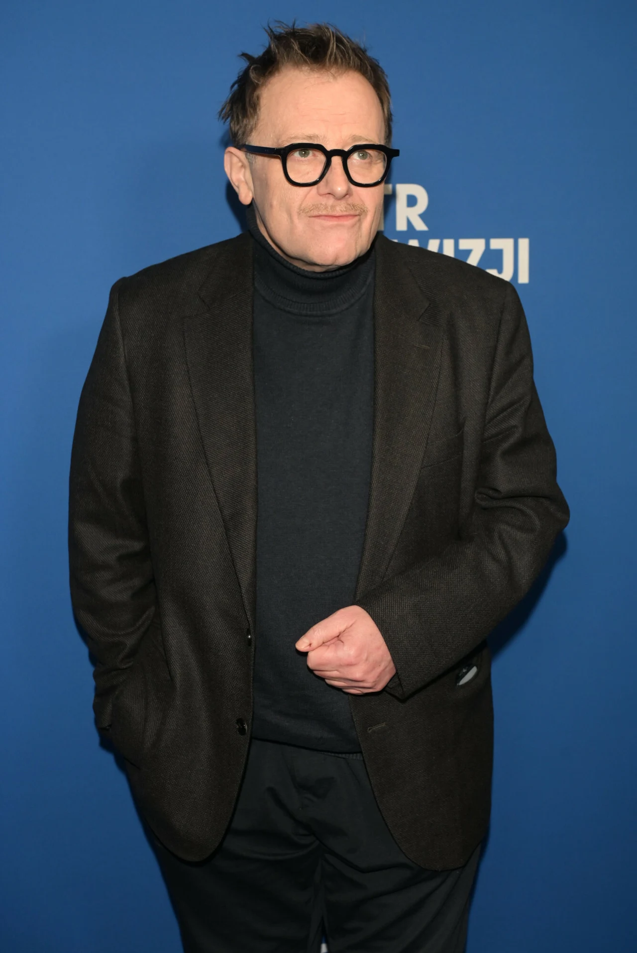 Mężczyzna w okularach, ubrany w czarną marynarkę i golf, stoi na tle niebieskiej ścianki z fragmentarycznie widocznym napisem.