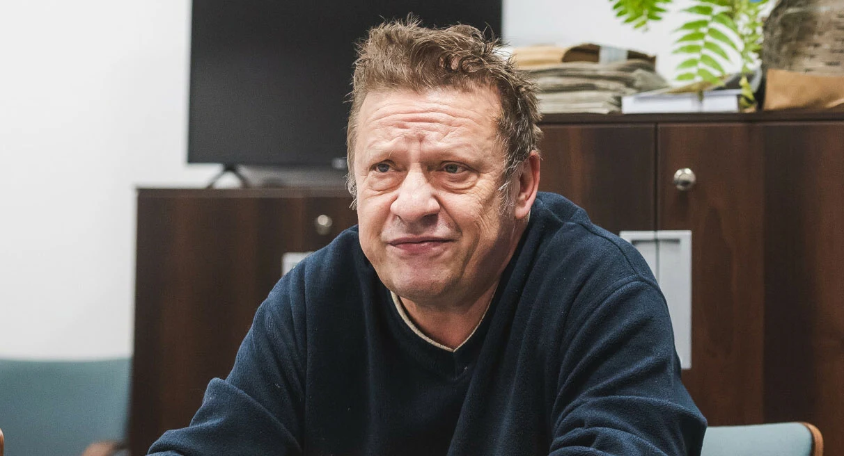 Jarek Janiszewski stoi na czele grupy Bielizna Mężczyzna w średnim wieku siedzi przy stole w biurze, ubrany w ciemny sweter, w tle brązowe szafki, stos dokumentów i zielona roślina.