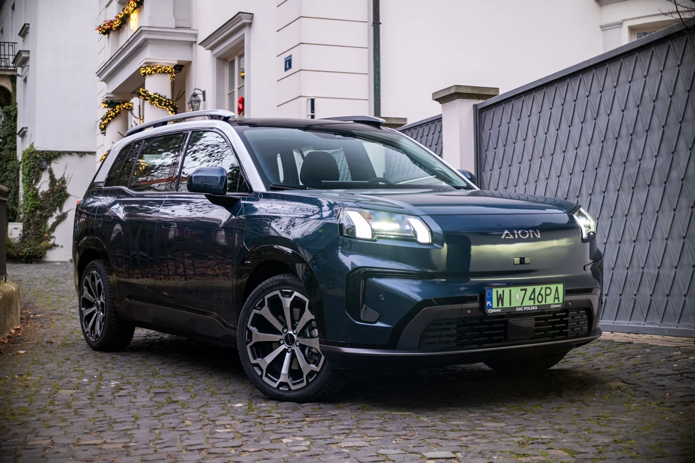 Ciemnozielony samochód elektryczny Aion SUV zaparkowany na brukowanej ulicy przy eleganckim budynku z ozdobami świątecznymi na balkonie, widoczne polskie tablice rejestracyjne.