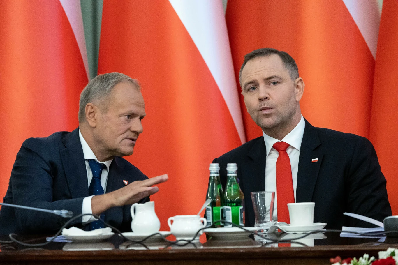 Prezydent i premier rozmawiali nt. uczestnictwa Polski w trumpowskiej Radzie Pokoju Dwóch mężczyzn w garniturach podczas oficjalnego spotkania, siedzących przy stole z filiżankami, szklankami i butelkami wody, na tle licznych flag Polski.