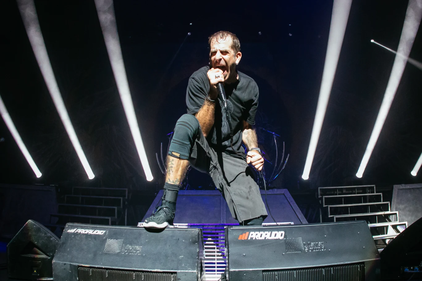 Randy Blythe (Lamb of God) w akcji Wokalista występujący na scenie podczas koncertu, intensywne światła sceniczne skierowane na niego, dynamiczna poza, mocno uchwycona ekspresja emocji, wokół sprzęt nagłaśniający oraz schody prowadzące na wyższy poziom sceny.
