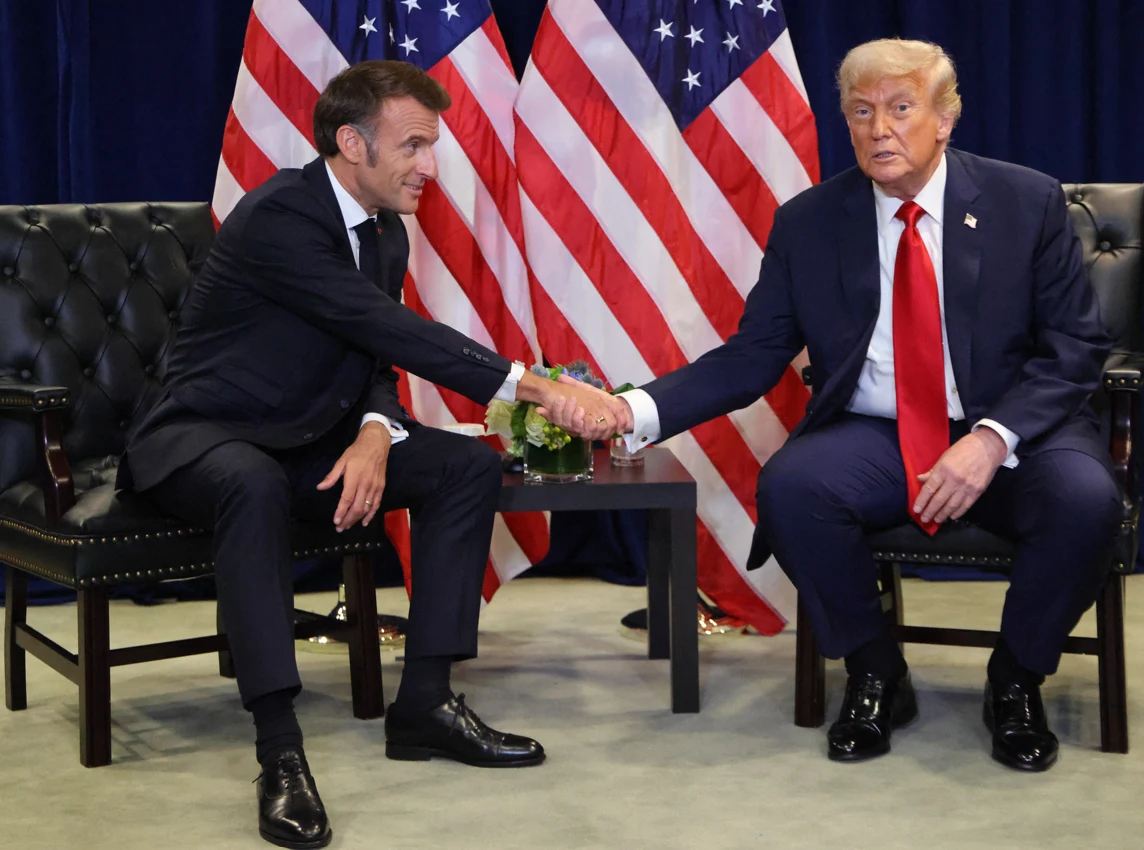 Dwóch mężczyzn w garniturach podaje sobie ręce podczas spotkania oficjalnego, siedząc naprzeciw siebie na czarnych fotelach na tle flag Stanów Zjednoczonych.