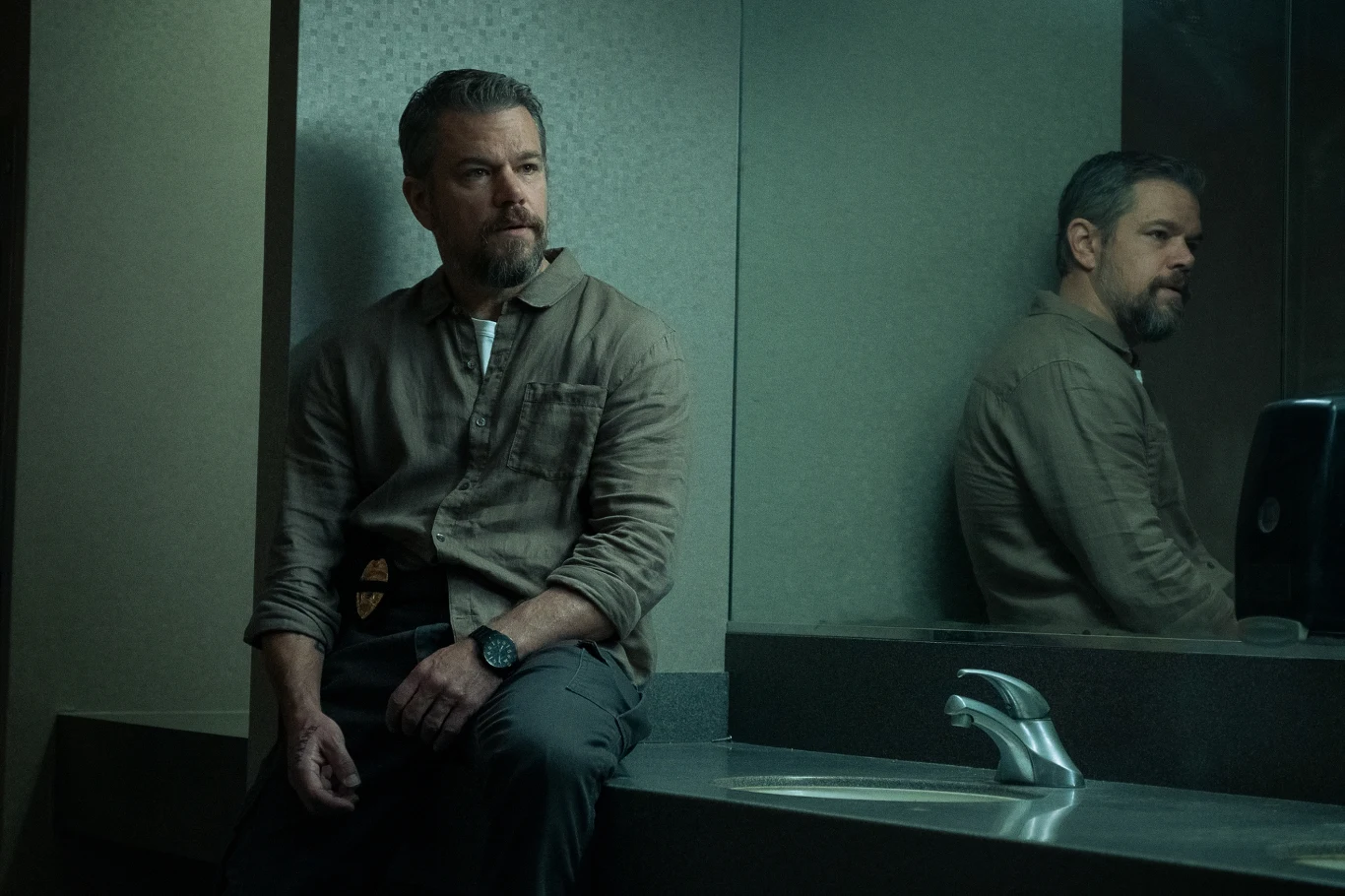 Mężczyzna o siwej brodzie siedzi przy umywalce w łazience, trzyma w ręku plakietkę identyfikacyjną, w tle widoczne jego odbicie w lustrze, jednolita kolorystyka ścian i oświetlenie nadają scenie poważny nastrój.