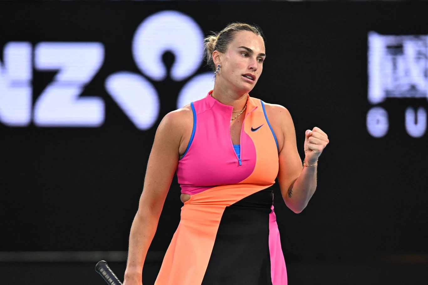 Tenisistka w sportowym stroju wykonuje gest triumfu z zaciśniętą pięścią, stojąc na korcie podczas meczu tenisowego. W tle widoczne logo sponsora.