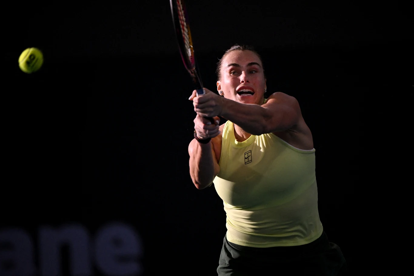 Tenisistka w jasnej koszulce wykonuje uderzenie dwuręcznym bekhendem, żółta piłka tenisowa widoczna w powietrzu, ciemne tło podkreśla dynamikę akcji.