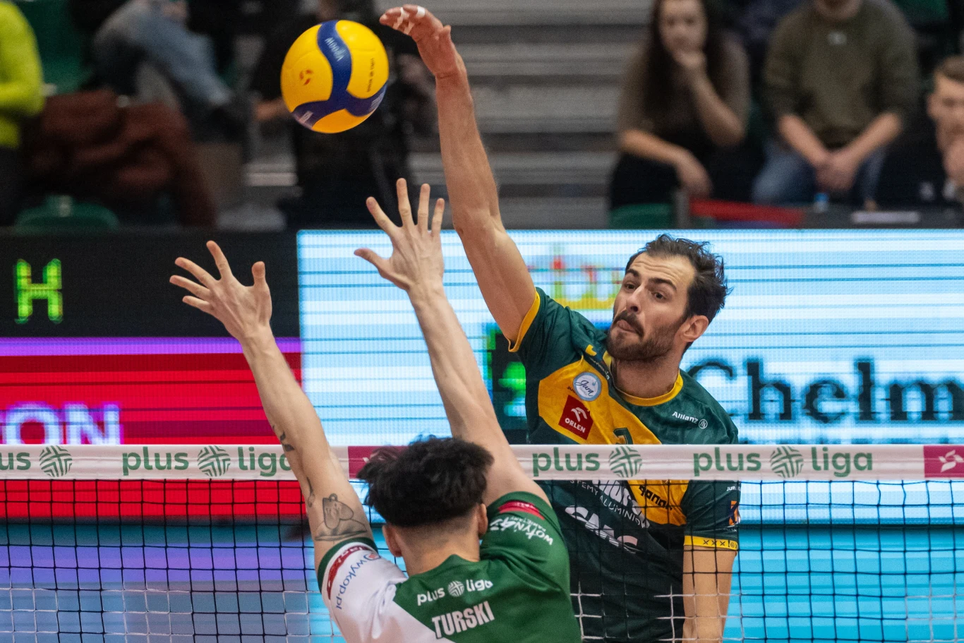 Zawodnik w zielono-żółtym stroju wykonuje atak siatkarski nad siatką, podczas gdy przeciwnik w zielono-białym stroju próbuje go zablokować, wokół widoczni kibice i elementy hali sportowej.