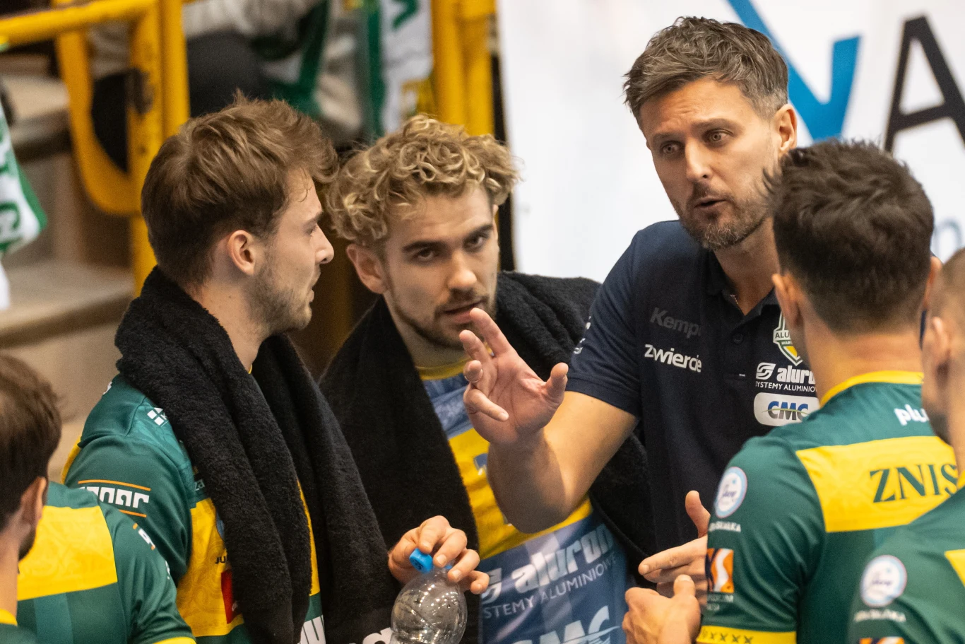 Trener drużyny siatkarskiej omawiający strategię z zawodnikami podczas przerwy w meczu, wszyscy skoncentrowani na rozmowie, sportowe stroje w barwach zespołu, na pierwszym planie wyraźny kontakt wzrokowy i gestykulacja.
