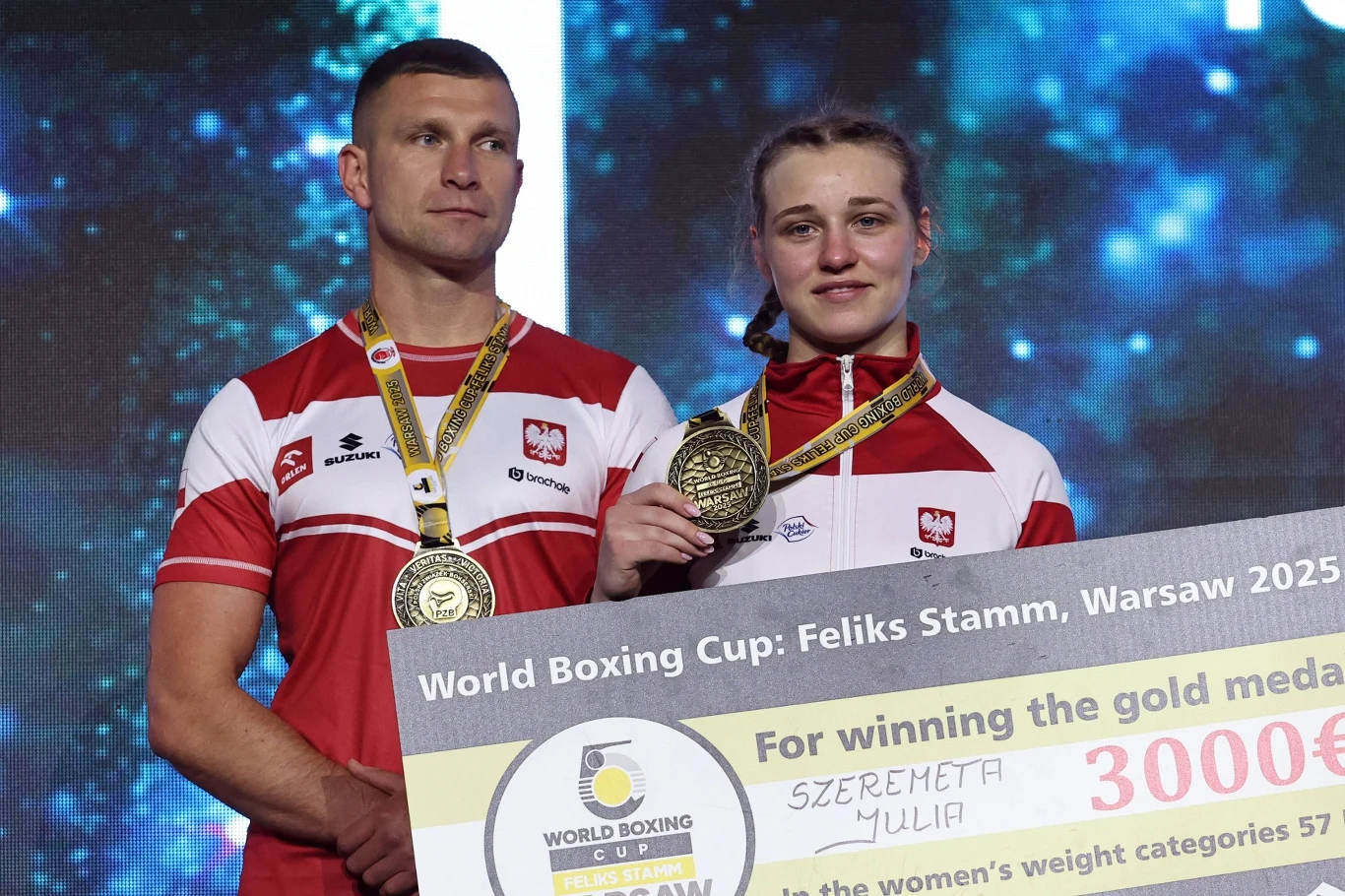 Dwoje sportowców w strojach reprezentacji Polski, noszących medale na szyjach, trzymających duży czek z nagrodą za zdobycie złotego medalu na międzynarodowym turnieju bokserskim w Warszawie.