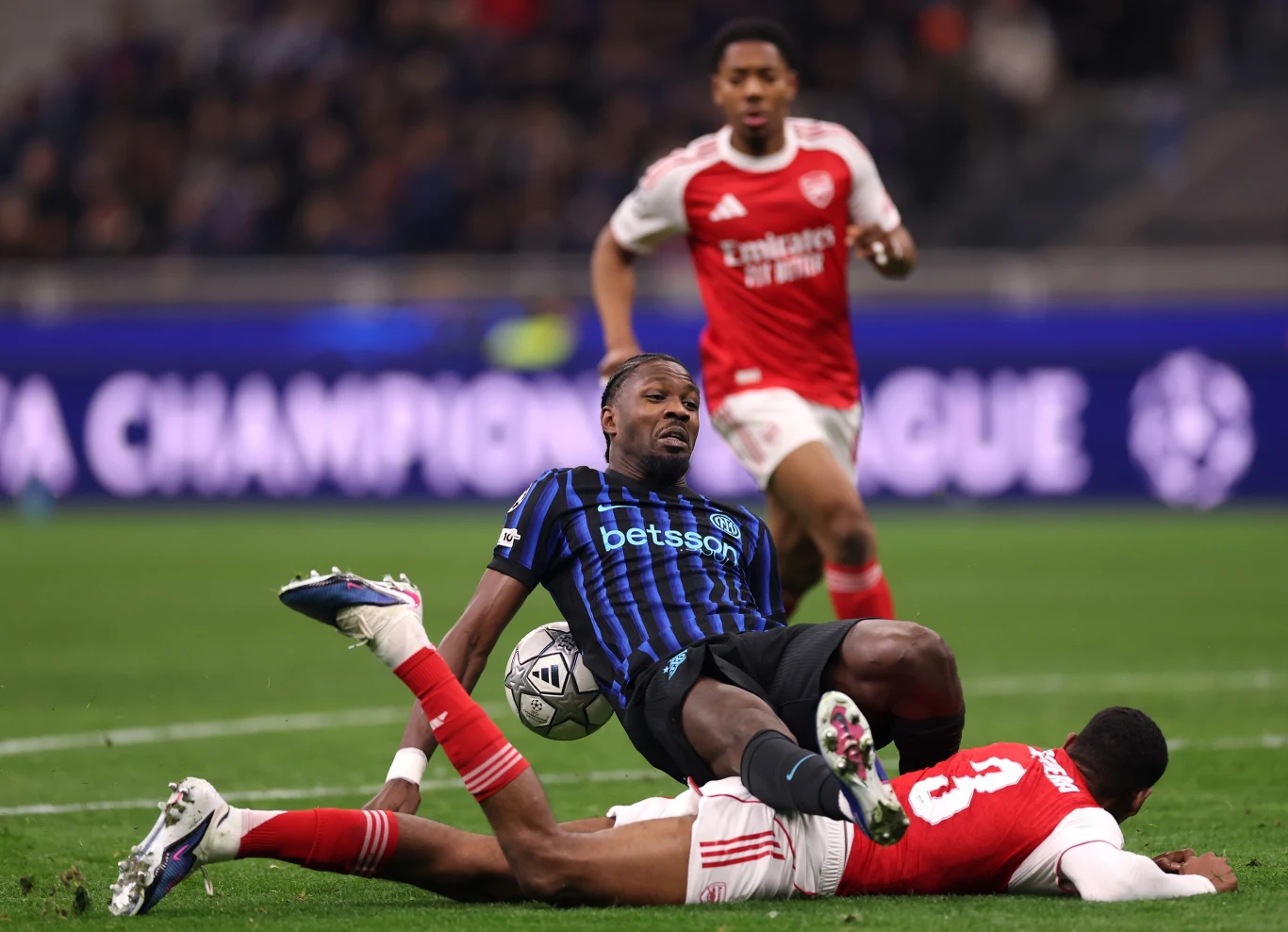 Zawodnik drużyny w niebiesko-czarnych pasach upada na murawę podczas dynamicznej walki o piłkę z rywalem w czerwono-białym stroju, w tle inny piłkarz obserwuje sytuację.