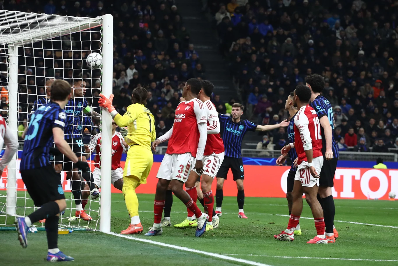 Zamieszanie w polu bramkowym podczas meczu piłki nożnej, bramkarz w żółtym stroju próbuje obronić piłkę, otoczony przez kilku zawodników jednej i drugiej drużyny, na tle wypełnionych trybun.