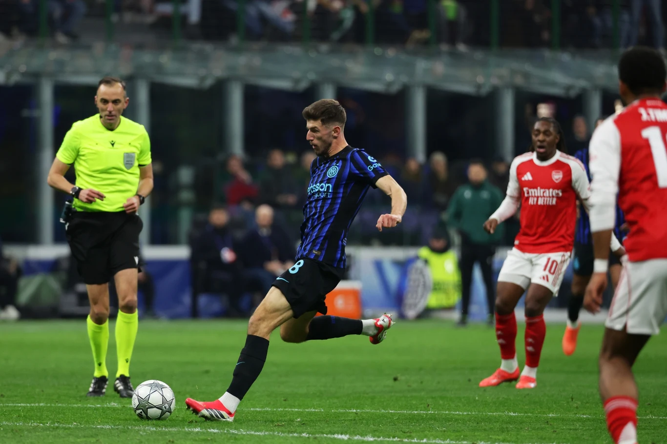 Piłkarz w czarno-niebieskim stroju kopie piłkę podczas meczu piłkarskiego, wokół widoczni są inni zawodnicy w czerwono-białych strojach oraz sędzia w żółtej koszulce, w tle kibice na trybunach.