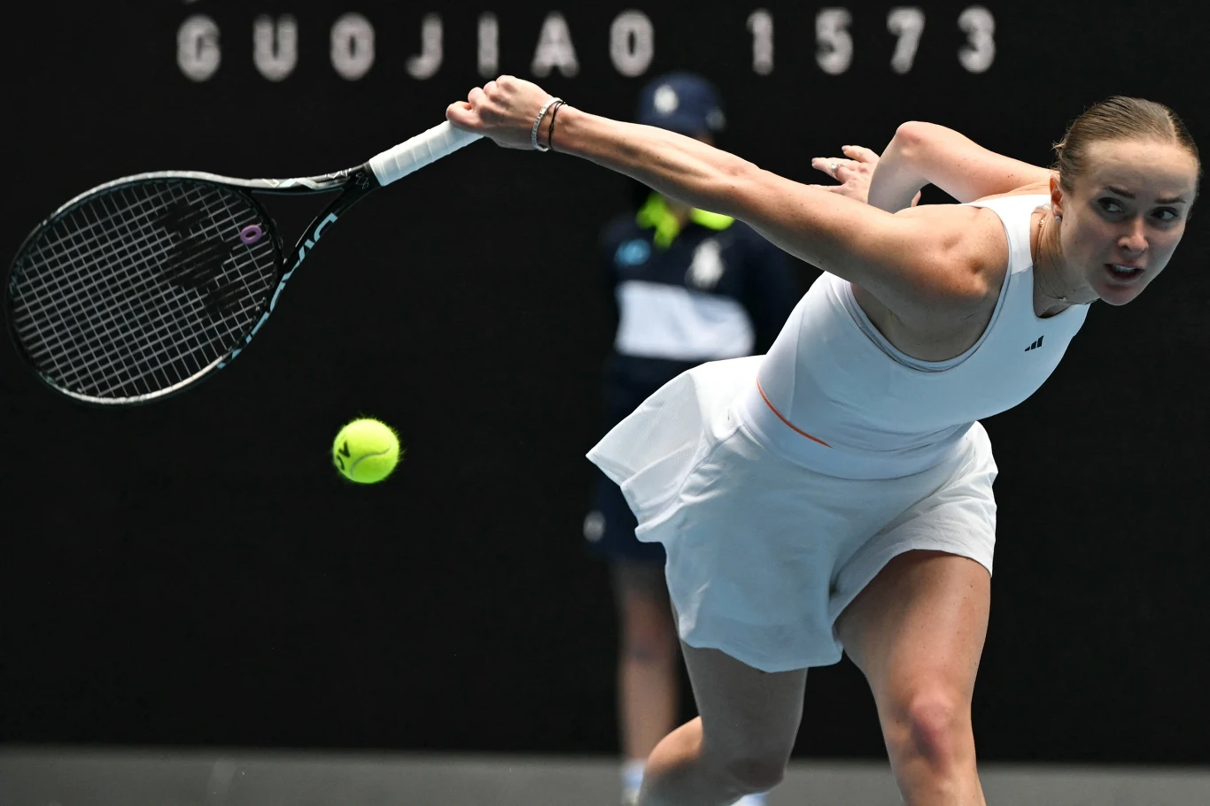 Tenisistka w białym stroju wykonuje dynamiczny zamach rakietą podczas gry, skupiona na żółtej piłce tenisowej, w tle widoczny sędzia lub asystent w ciemnym stroju.