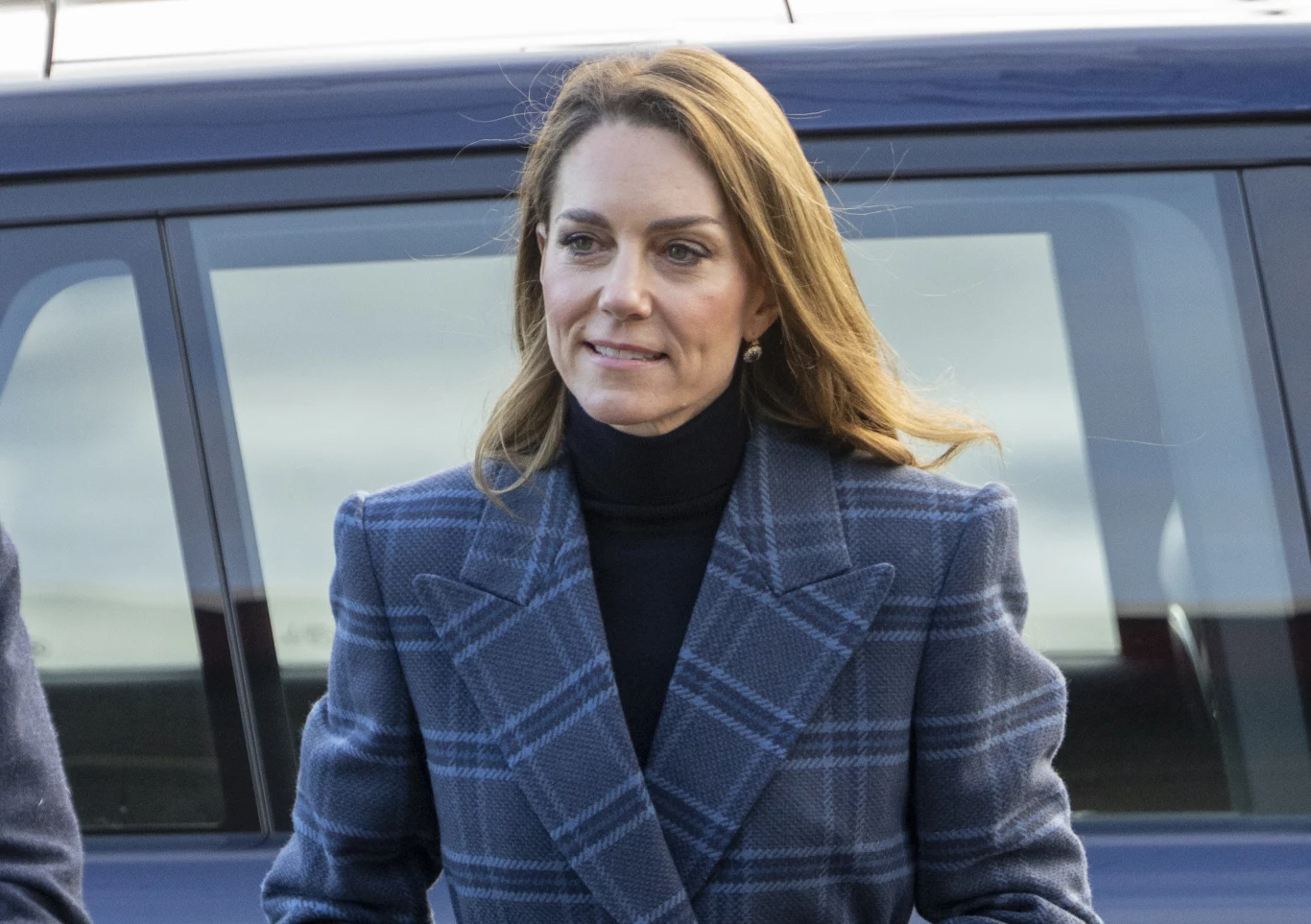 Księżna Kate udała się z wizytą do Szkocji Kobieta w eleganckim płaszczu w kratę i czarnym golfie stoi na tle samochodu, lekko uśmiechnięta, patrząc w bok.
