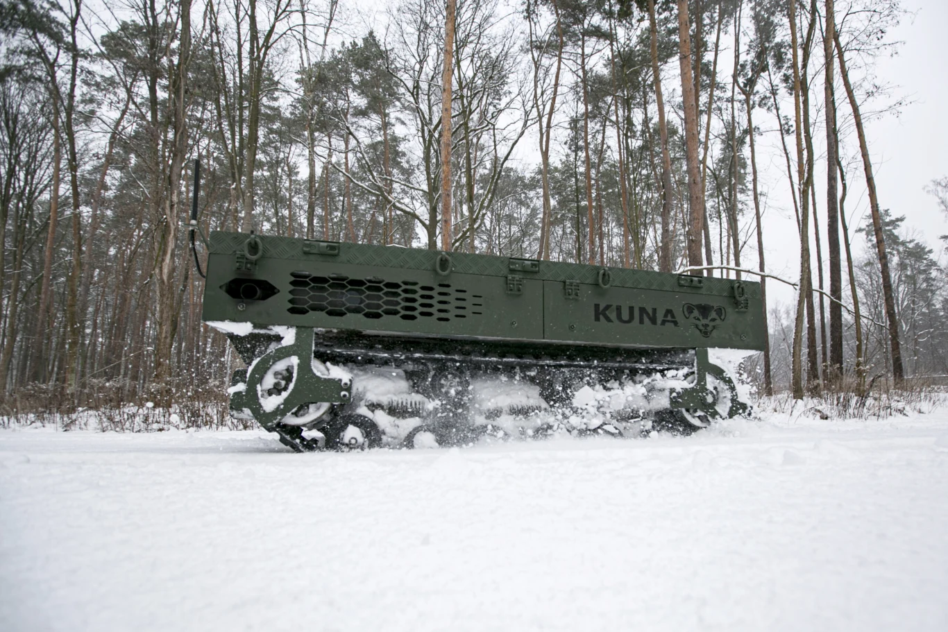 Bezzałogowy pojazd wojskowy o nazwie KUNA poruszający się po ośnieżonym lesie, otoczony wysokimi, bezlistnymi drzewami sosnowymi, wyraźne ślady na śniegu pod gąsienicami.