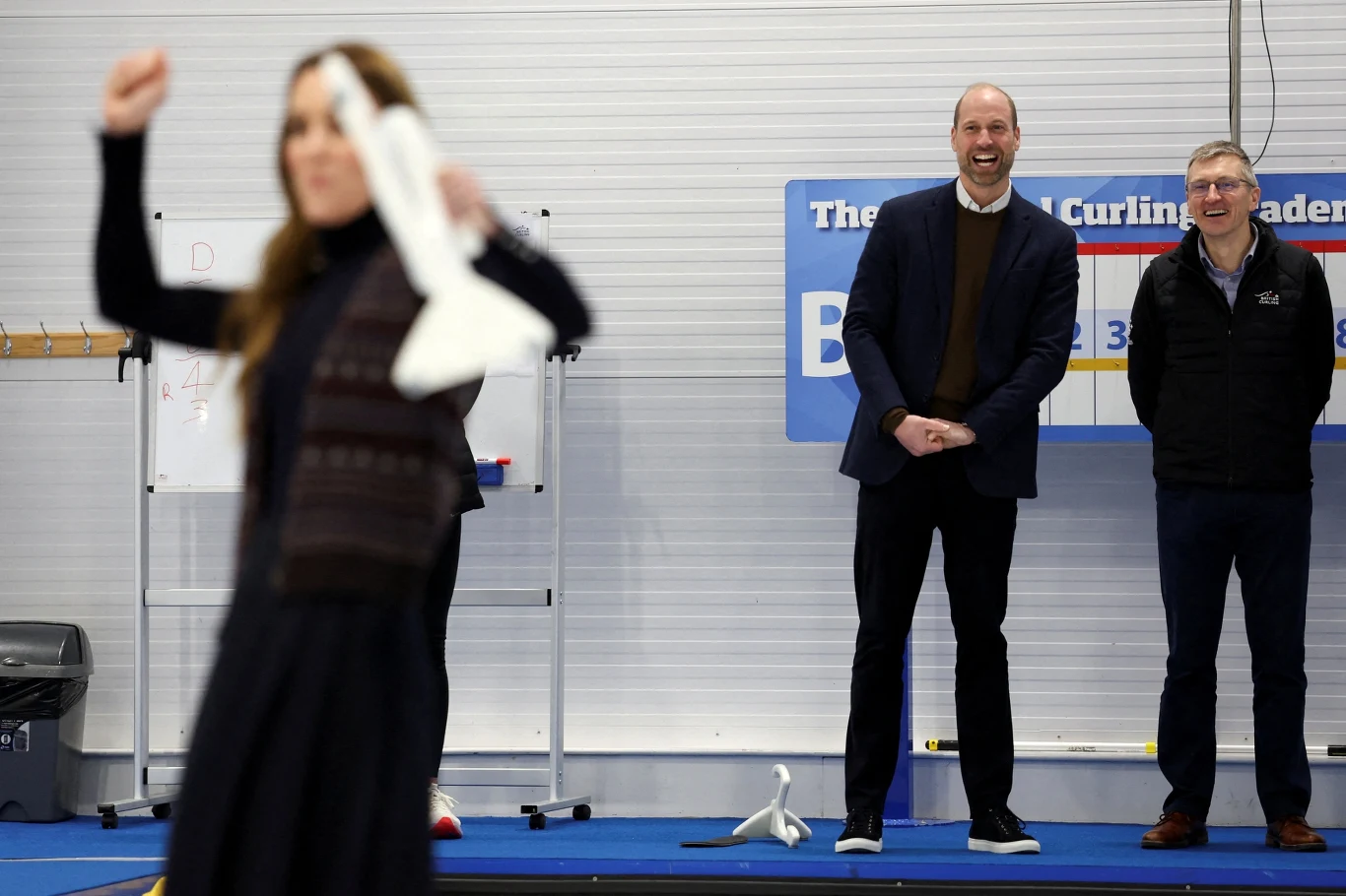 Książę William i księżna Kate zagrali w curling Trzy osoby biorą udział w zabawie lub ćwiczeniu z papierowymi samolotami w pomieszczeniu sportowym, na pierwszym planie kobieta w ruchu, w tle dwóch mężczyzn obserwujących sytuację z uśmiechem.