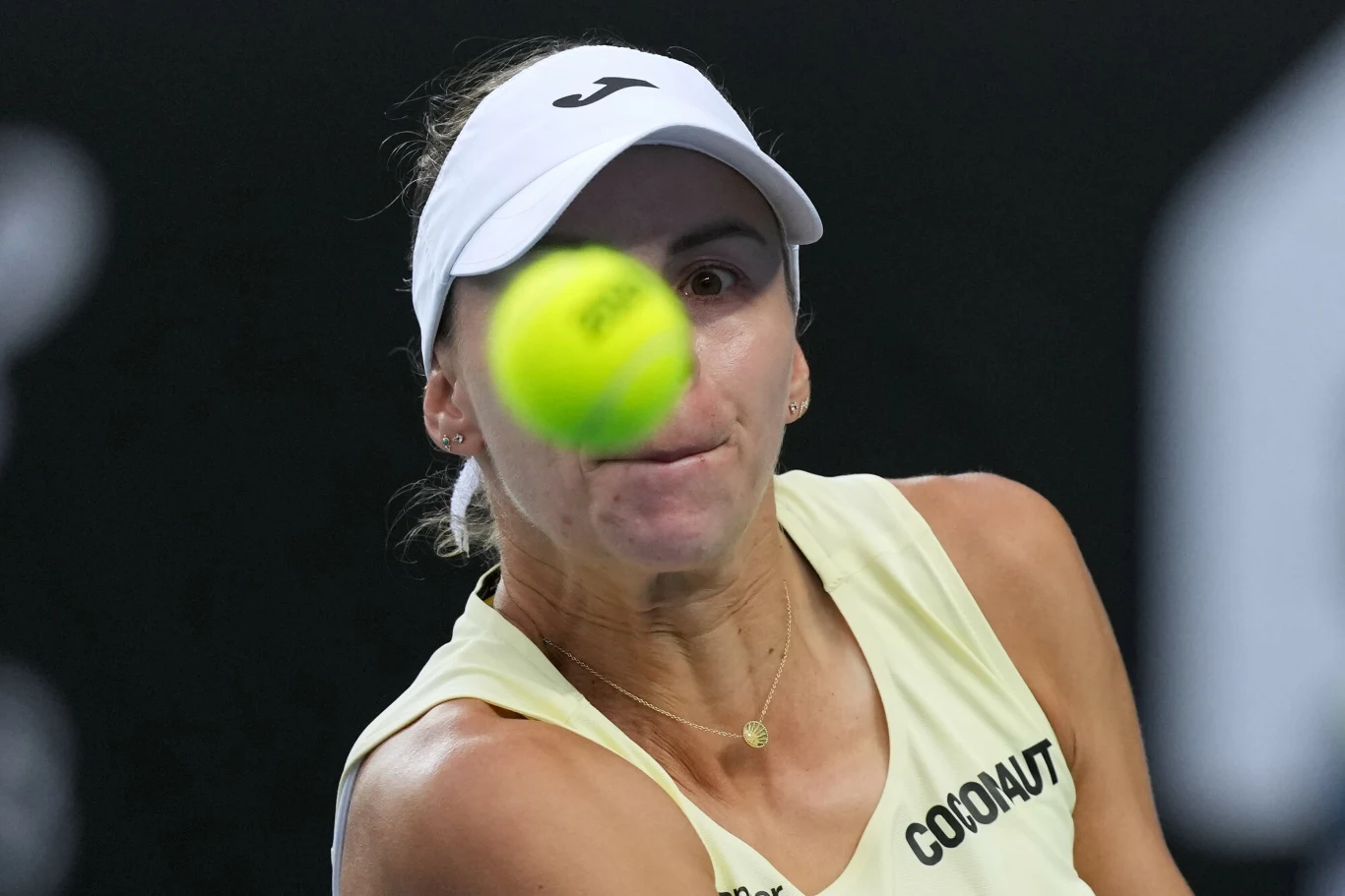 Zawodniczka tenisowa koncentruje wzrok na zbliżającej się piłce tenisowej, mając na sobie jasny strój sportowy i daszek, a tło jest ciemne i rozmazane.