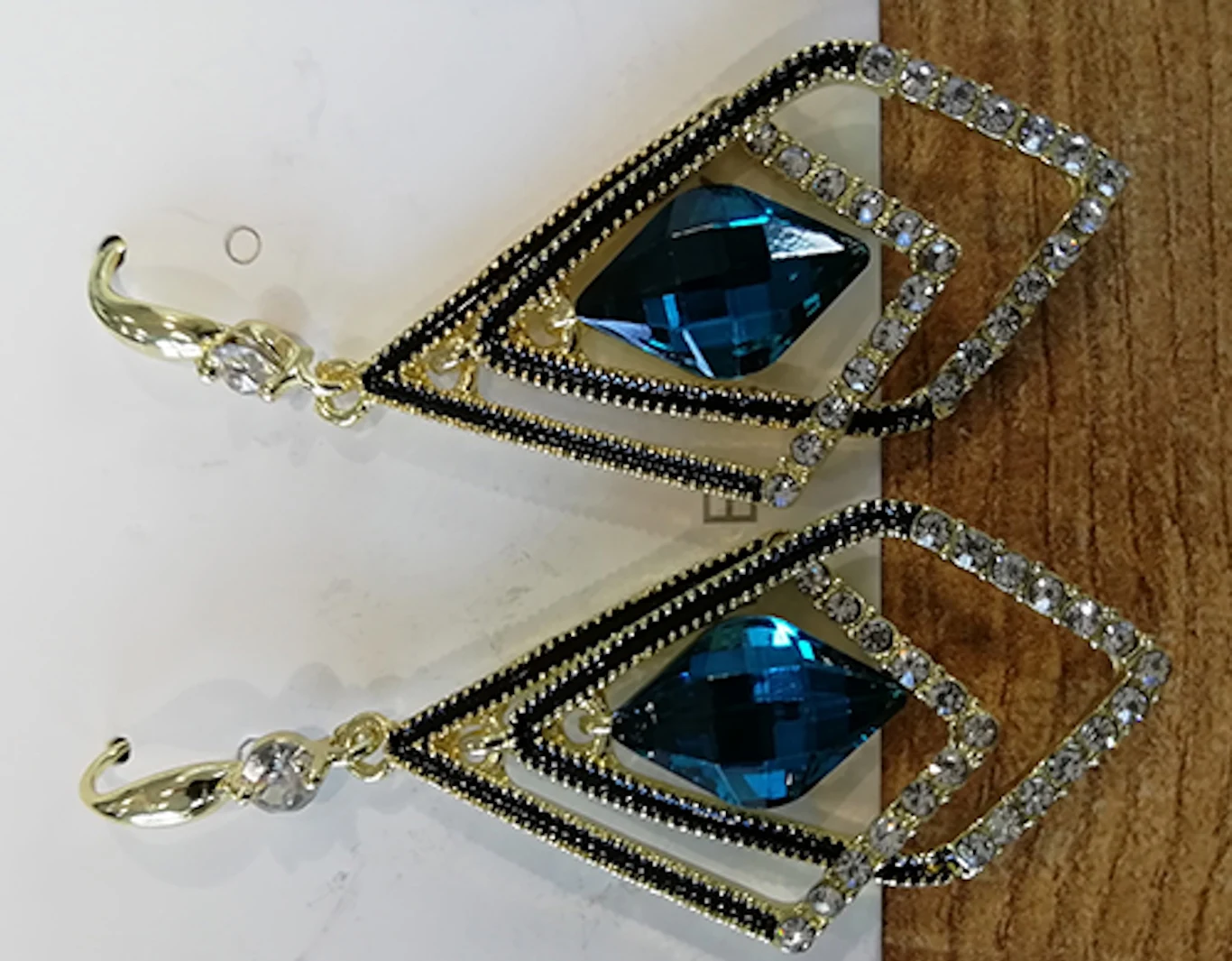 Para wiszących kolczyków o geometrycznym kształcie z ozdobnymi kryształami w kolorze niebieskim otoczonymi rzędami drobnych białych kamieni, na złotych zapięciach, ułożone na tle w odcieniu drewna i bieli.