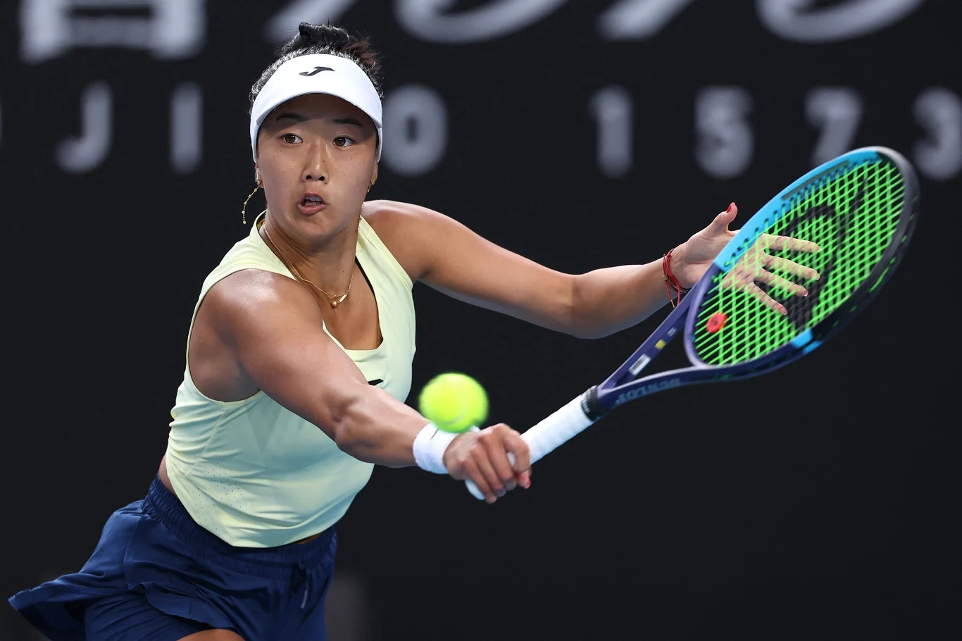 Tenisistka uderzająca piłkę rakietą podczas meczu tenisowego, skupiona twarz, dynamiczne ujęcie z widoczną piłką w locie, sportowa odzież i tło kortu tenisowego.