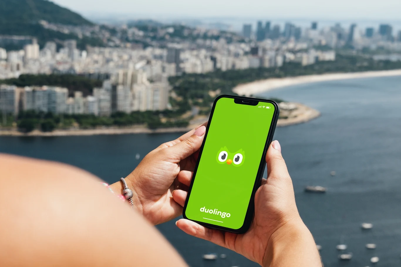 Dwa tygodnie testów Super Duolingo. Czy opłata się wykupić plan? Telefon z zielonym ekranem aplikacji Duolingo trzymany w dłoniach nad panoramą miasta i wybrzeża, w tle wysokie budynki i szerokie wody zatoki.