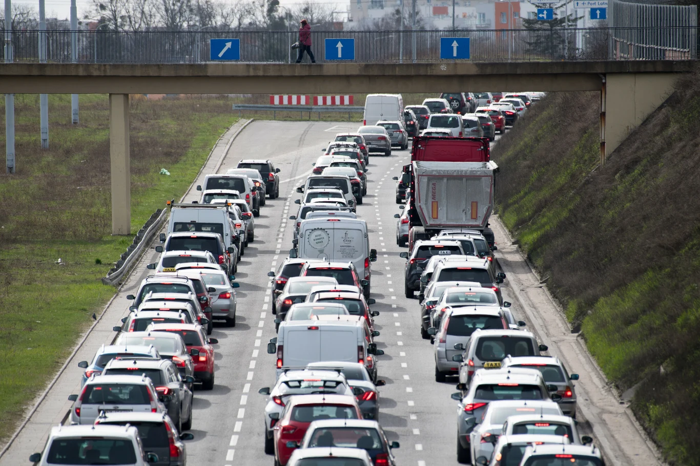 Korek drogowy na dwupasmowej ulicy w mieście, liczne samochody zatrzymane na całej szerokości jezdni, nad jezdnią kładka dla pieszych z jedną osobą.