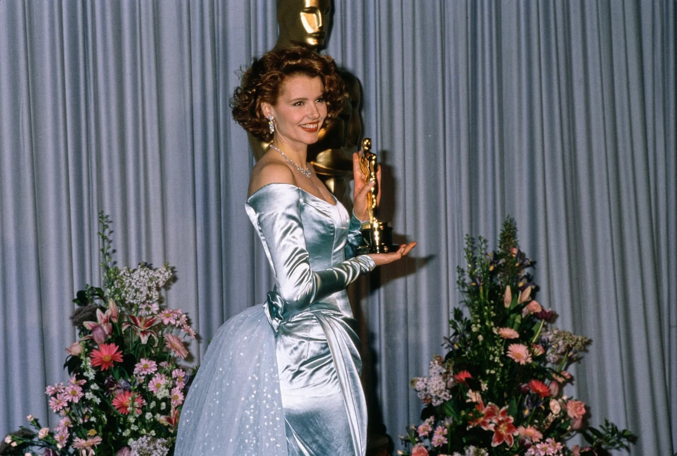Geena Davis z Oscarem za drugoplanową rolę w "Przypadkowym turyście" Elegancko ubrana kobieta w długiej jasnej sukni uśmiecha się, trzymając w dłoni statuetkę Oscara, w tle znajduje się duża statua Oscara oraz dekoracje z kwiatów i zasłonka.