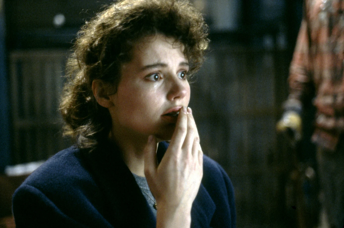 Geena Davis w filmie "Mucha" Młoda kobieta z wyrazem zmartwienia i niepokoju na twarzy, trzyma rękę przy ustach, w tle widoczne rozmyte postacie i metalowe pręty.