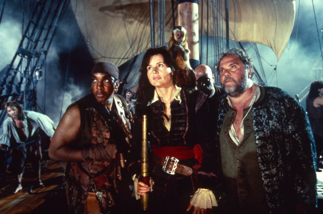 Kadr z filmu "Wyspa piratów" Trzech ubranych w pirackie stroje ludzi stoi na pokładzie statku żaglowego, z żaglem rozpostartym w tle i postaciami drugoplanowymi przygotowującymi się do podróży lub bitwy. Centralna postać trzyma lunetę.