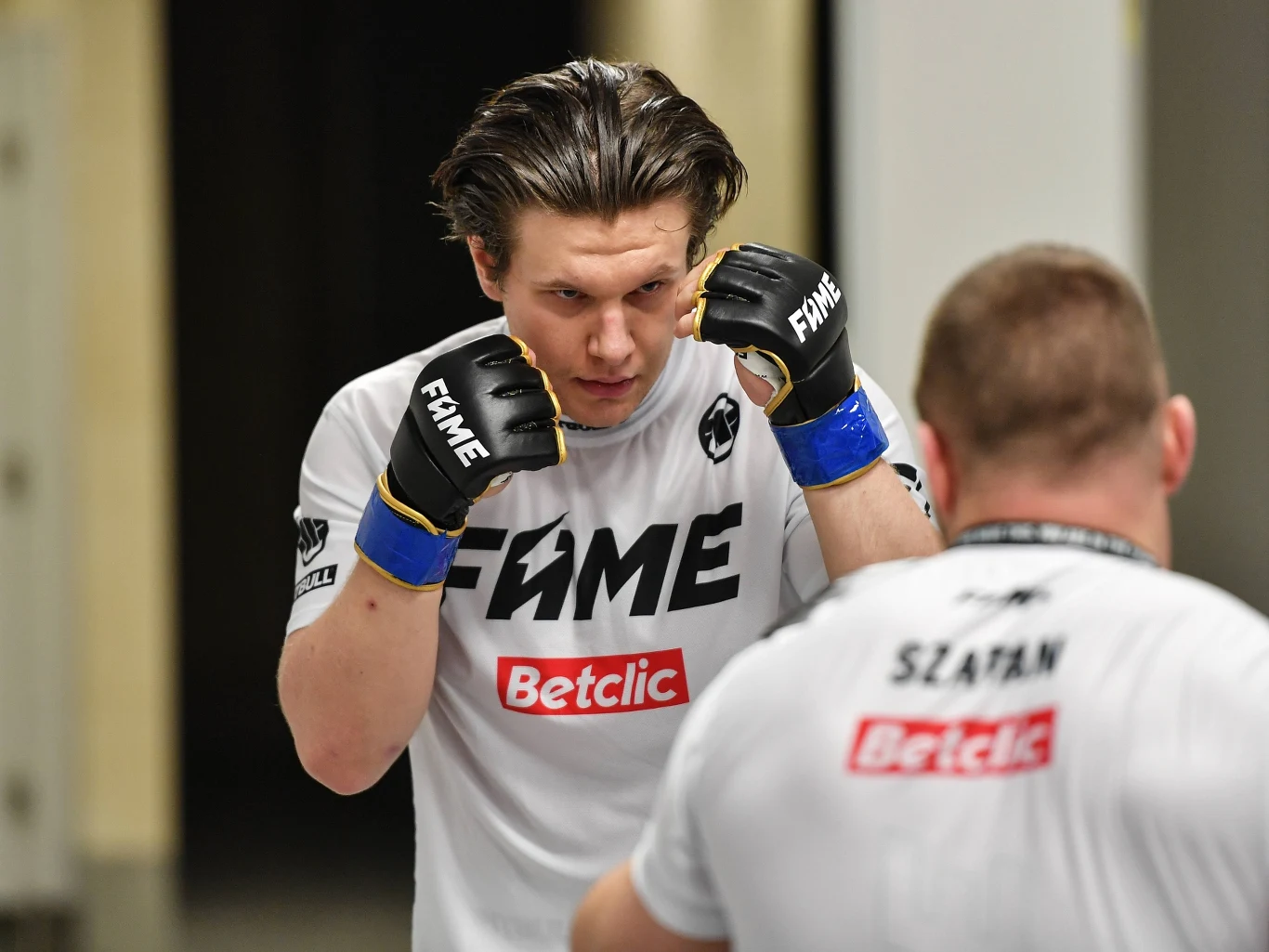 Natan Marcoń Dwóch zawodników w białych koszulkach z logo FAME i Betclic przygotowuje się do walki, jeden z nich stoi naprzeciw drugiego w gardzie z uniesionymi rękoma i skupionym wyrazem twarzy. Tło rozmyte, cała uwaga skupiona na sportowcach.