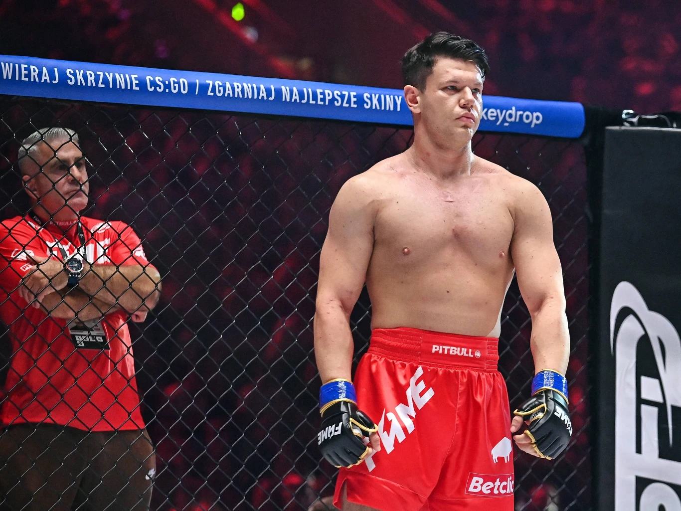 Natan Marcoń Zawodnik MMA w czerwonych spodenkach i rękawicach stoi w oktagonie w oczekiwaniu na walkę, za nim znajduje się sędzia lub członek obsługi technicznej ubrany w czerwoną koszulkę.