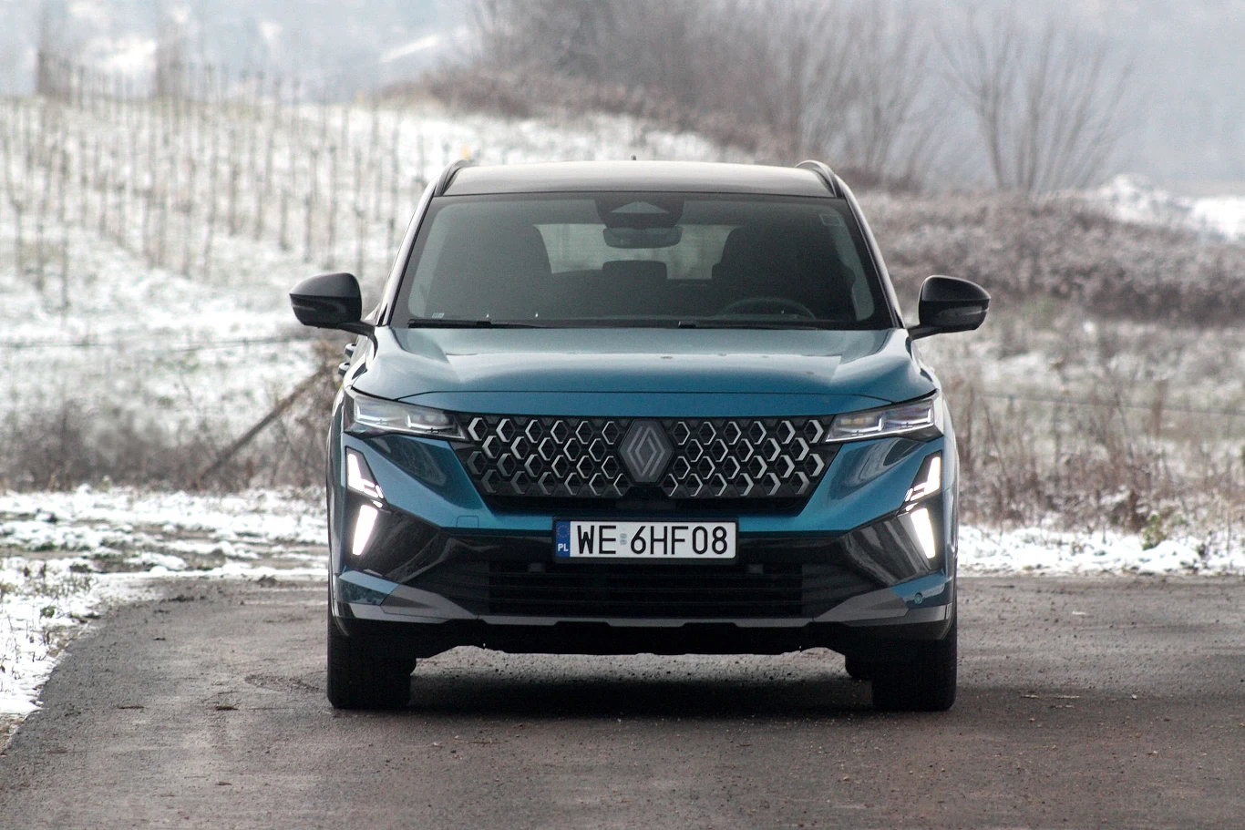 Niebieski SUV marki Renault stoi zaparkowany na drodze asfaltowej, wokół teren lekko pokryty śniegiem, w tle widoczna roślinność i pagórkowaty krajobraz.