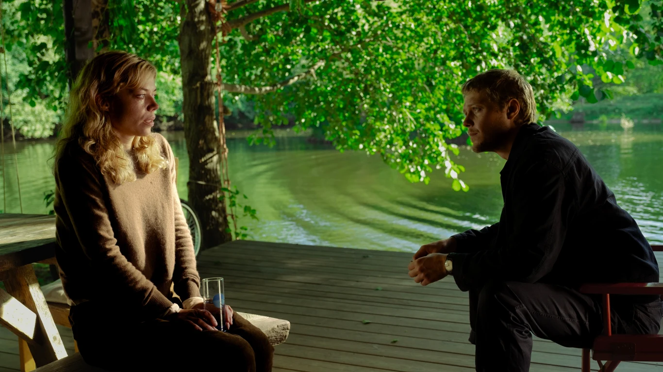 Dwie osoby siedzą na drewnianym tarasie nad jeziorem, otoczone zielonymi drzewami. Kobieta trzyma szklankę, a naprzeciwko niej mężczyzna w zamyślonej pozie.
