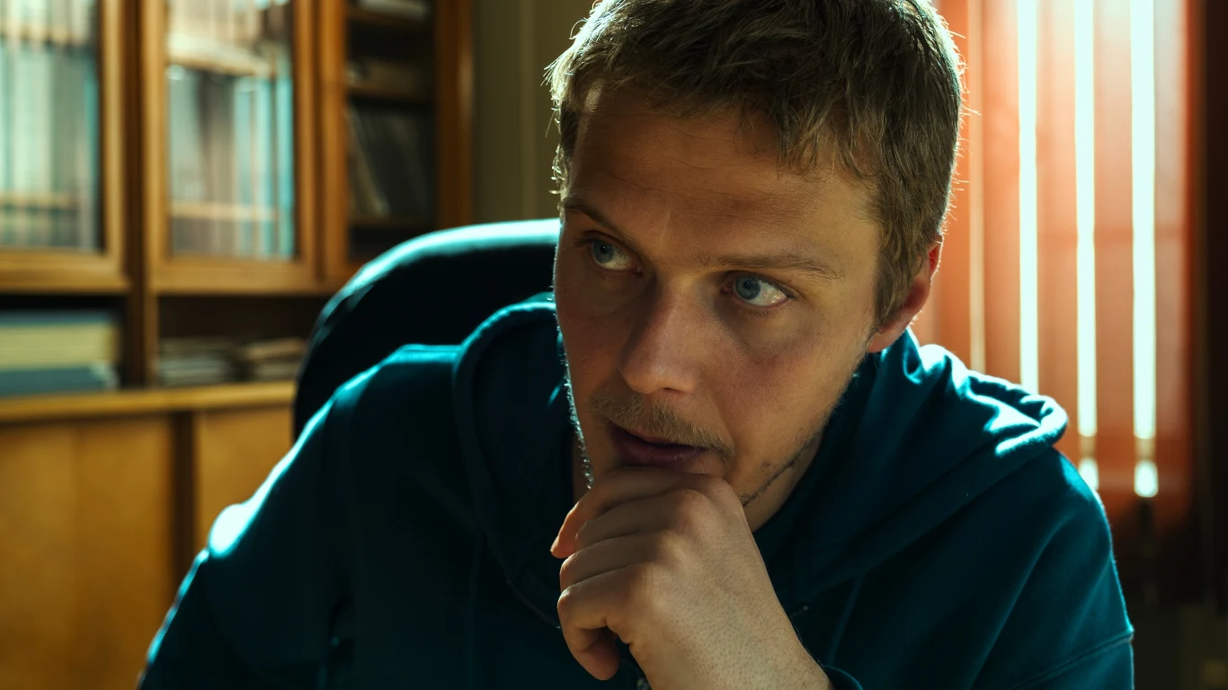 Młody mężczyzna w błękitnej bluzie siedzi przy biurku, z twarzą opartą na dłoni, wpatrzony w coś z wyrazem zamyślenia. W tle regał z książkami oraz promienie światła wpadające przez żaluzje.