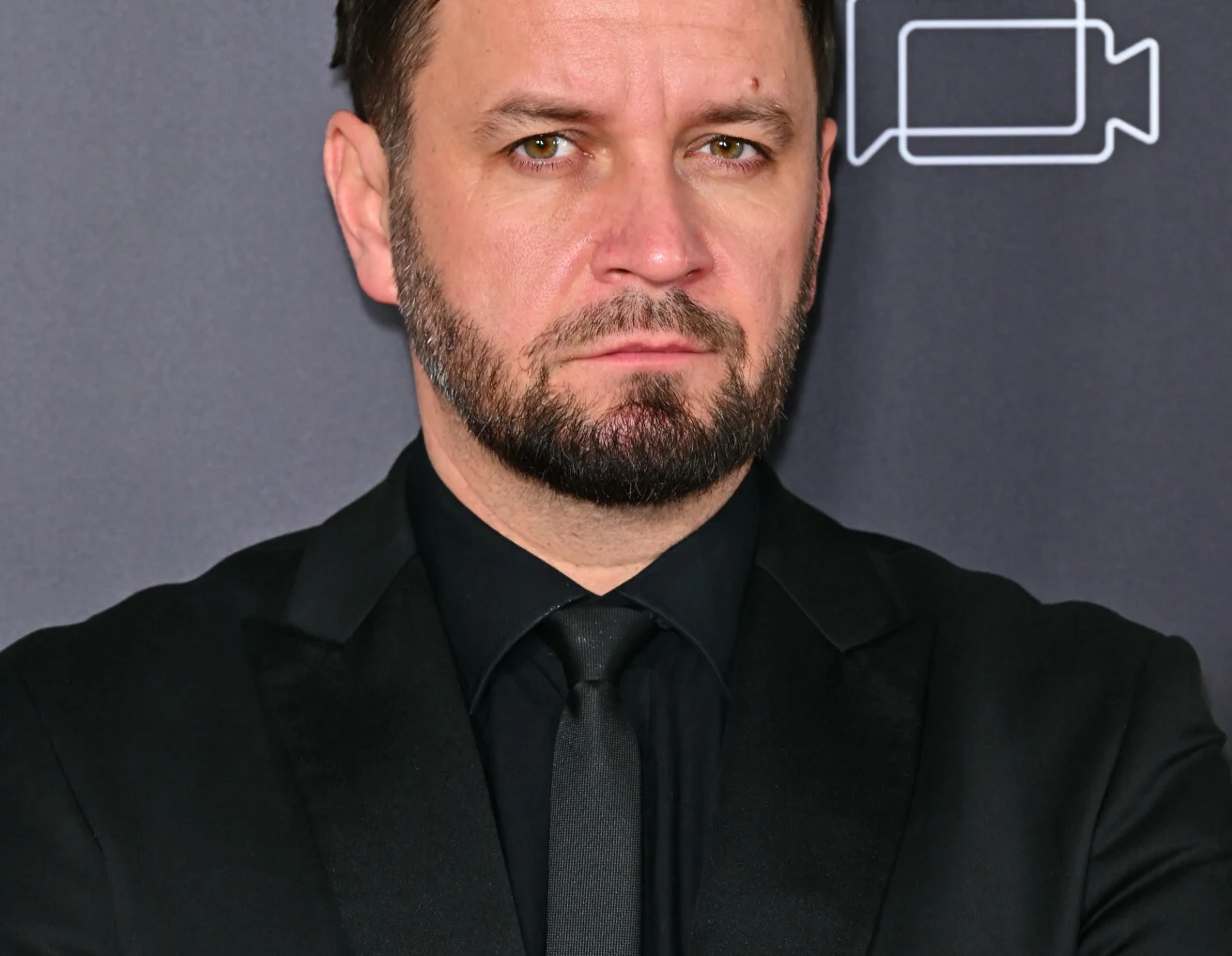 Piotr Domalewski Mężczyzna z brodą ubrany w czarną marynarkę, czarną koszulę i czarny krawat na tle grafiki z symbolem kamery.