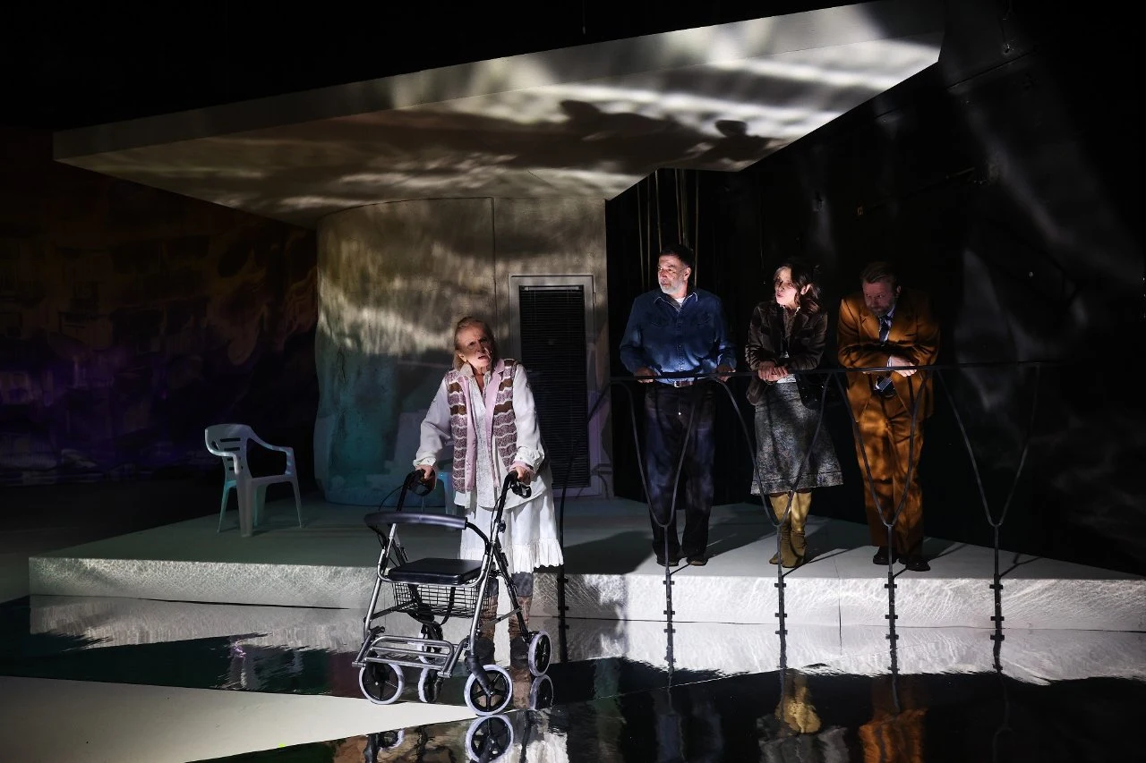 Kadr ze spektaklu "Zamknij oczy Nel. W pustyni i w puszczy", fot. Andrzej Banas / Narodowy Stary Teatr w Krakowie Czworo aktorów na scenie teatralnej, jedna kobieta stoi z chodzikiem na tle nowoczesnej scenografii, pozostali trzymają się barierki, światło tworzy dynamiczne refleksy na tle.