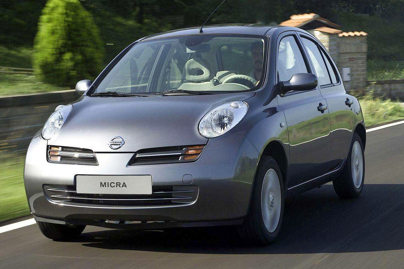 Srebrny samochód osobowy Nissan Micra porusza się po asfaltowej drodze, otoczony zielenią i elementami przydrożnej architektury, w tle widoczne jest niskie ogrodzenie oraz fragmenty gęstych krzewów.