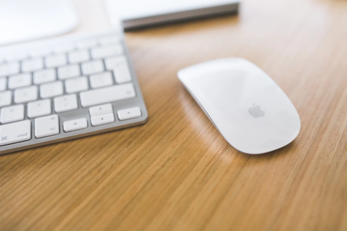 Biała myszka Apple typu Magic Mouse oraz klawiatura komputerowa na drewnianym biurku, w tle fragment laptopa.
