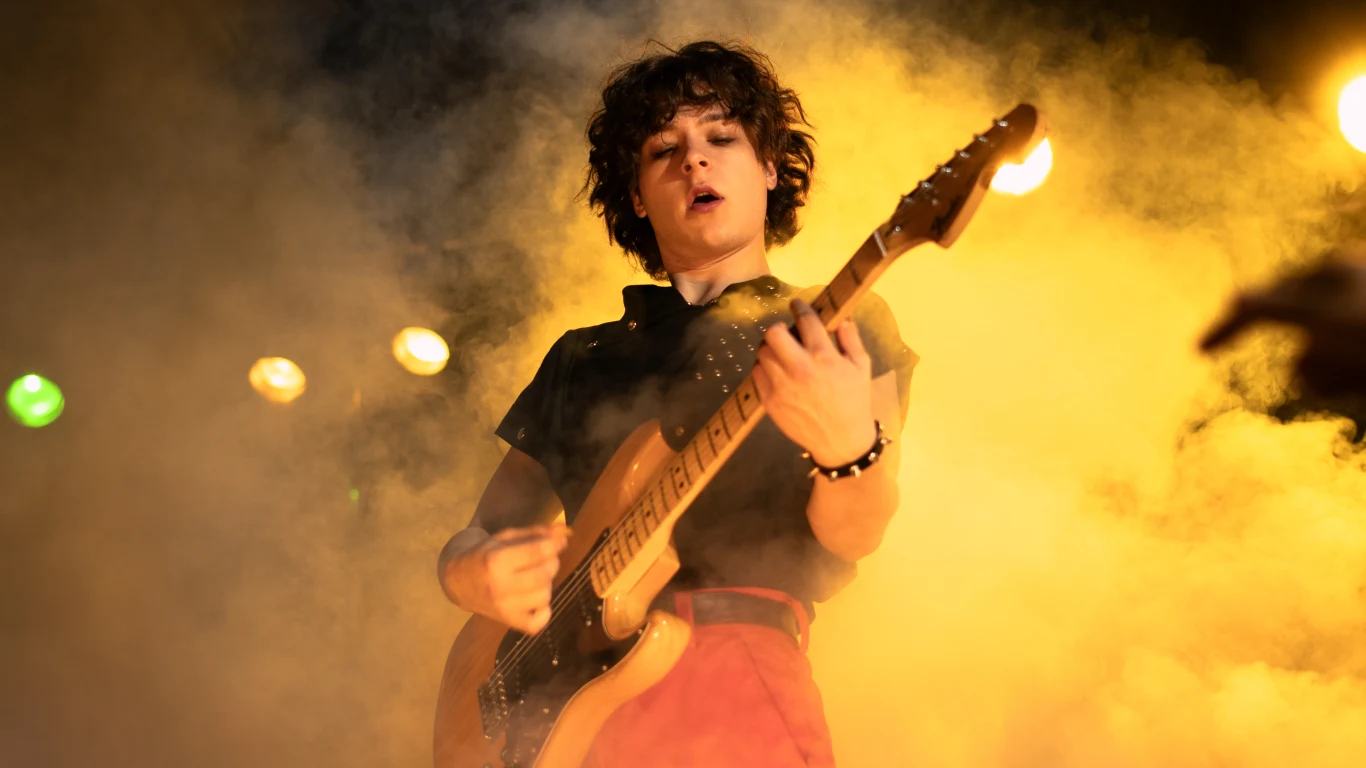 Młody muzyk grający na gitarze elektrycznej w otoczeniu intensywnych świateł scenicznych i dymu, co tworzy dynamiczny i energetyczny klimat koncertu.
