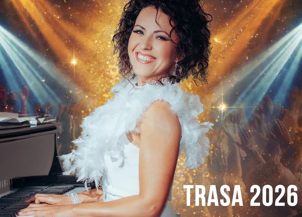 Hania Stach Uśmiechnięta kobieta z kręconymi ciemnymi włosami w eleganckiej białej sukni gra na fortepianie, za nią widać sceniczne światła i rozświetlone tło oraz napis Trasa 2026.