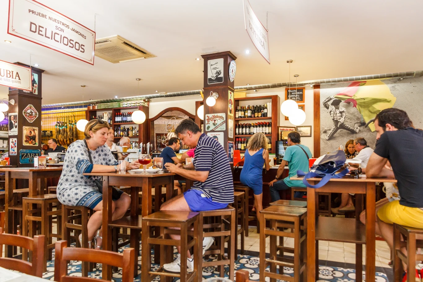 W Hiszpani napiwek nie jest obowiązkowy. Goście siedzący przy drewnianych stołach zajęci rozmową i jedzeniem w kolorowej, przytulnej restauracji; na ścianach widoczne dekoracje, plakaty i półki z winami.