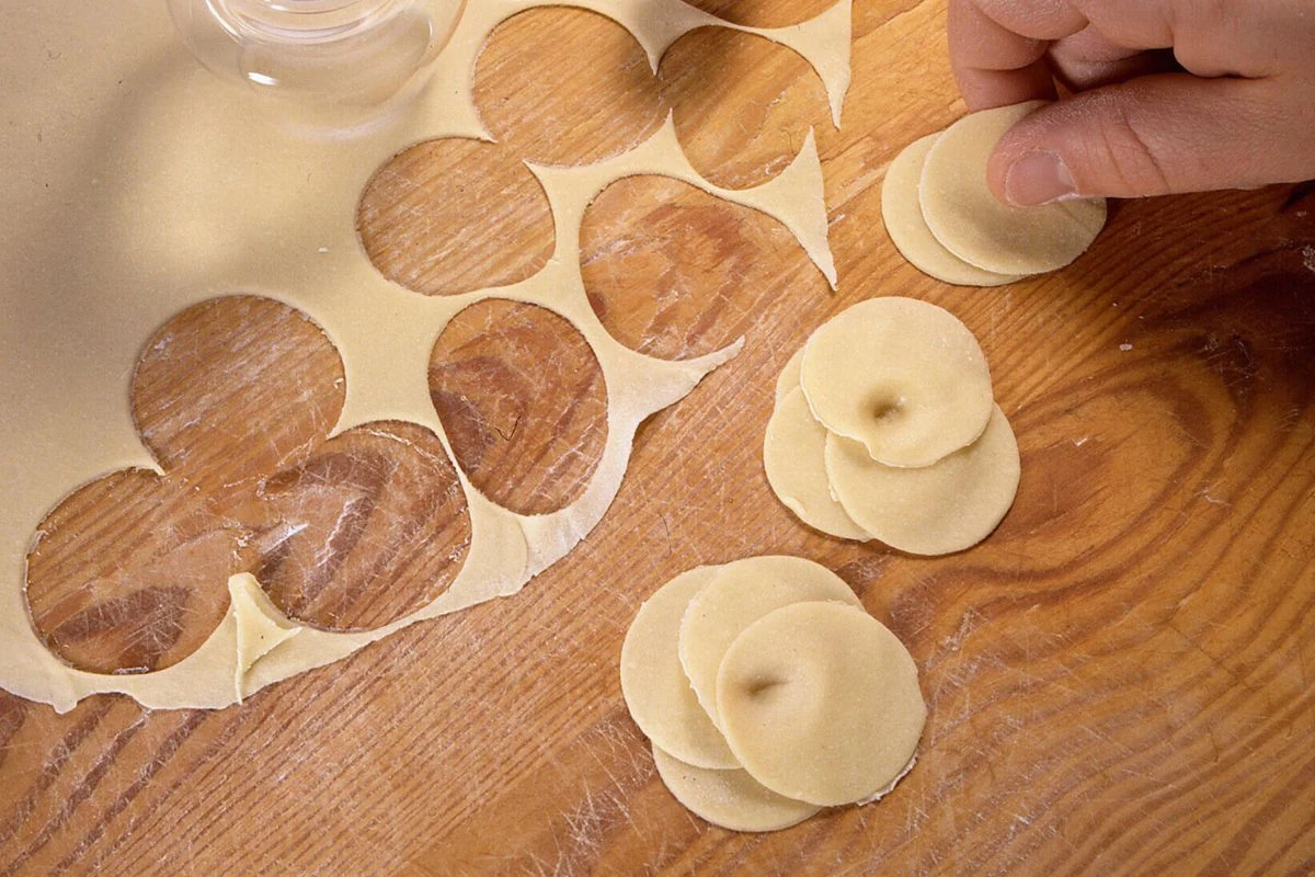 Ręka układająca okrągłe kawałki ciasta na drewnianym blacie, ciasto zostało wykrojone specjalną foremką, część wykrojonych już placków ułożona jest w małe stosiki obok rozwałkowanego ciasta.