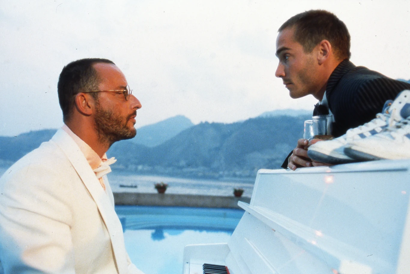 Dwóch mężczyzn rozmawia przy białym fortepianie ustawionym na tle basenu i gór, jeden z nich ubrany jest w biały garnitur, drugi opiera się o fortepian trzymając szklankę, atmosfera rozmowy jest poważna i skupiona.